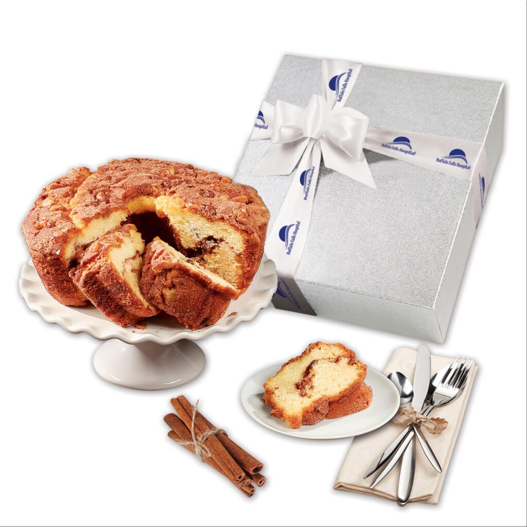 A delicious cinnamon coffee cake is pictured on a cake stand with a slice on a plate next to it. Behind the cake is a silver box tied with a custom branded ribbon.