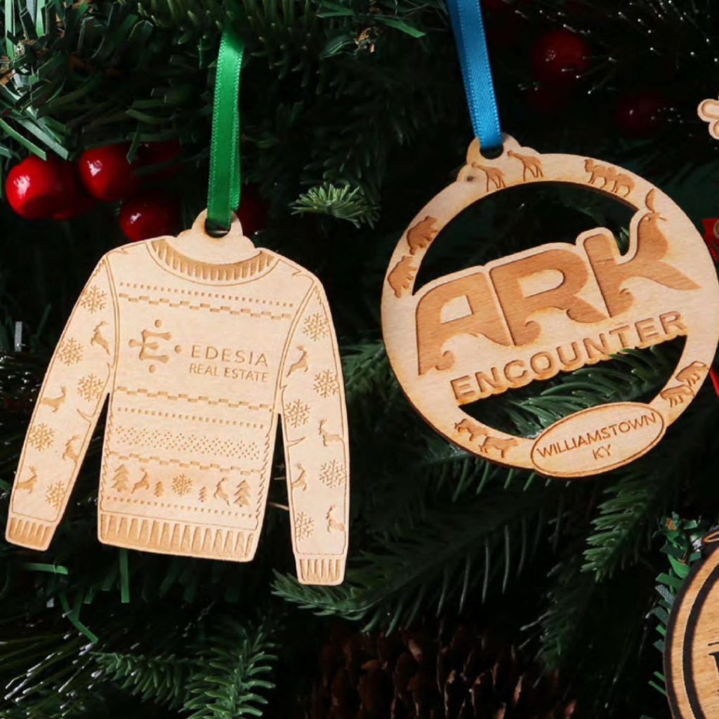 Two wooden ornaments hang from a tree. One is an ugly sweater and the other is a round ornament
