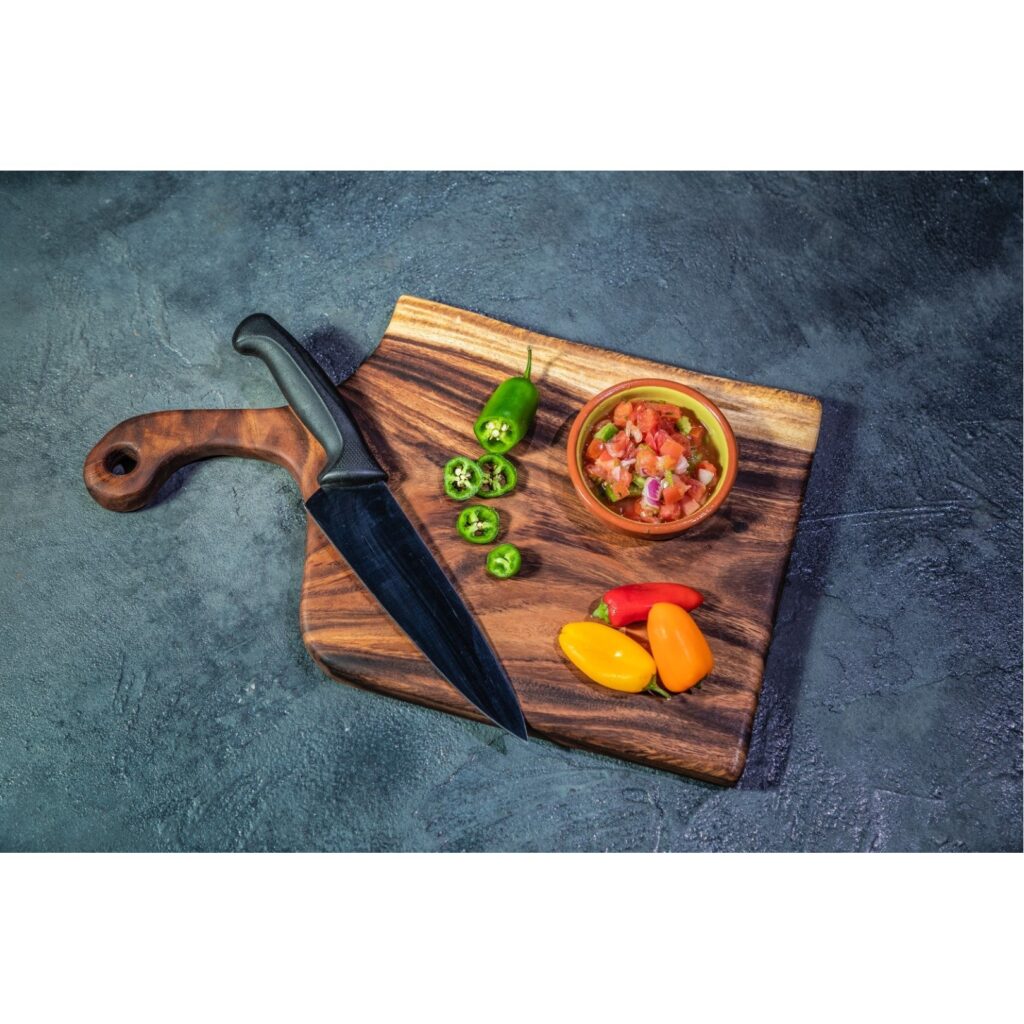A wooden cutting board lays on a blue surface. The board isn't perfectly squared, it curves on a few sides and the handle is curved as well. Vegetables and a knife sit on the board