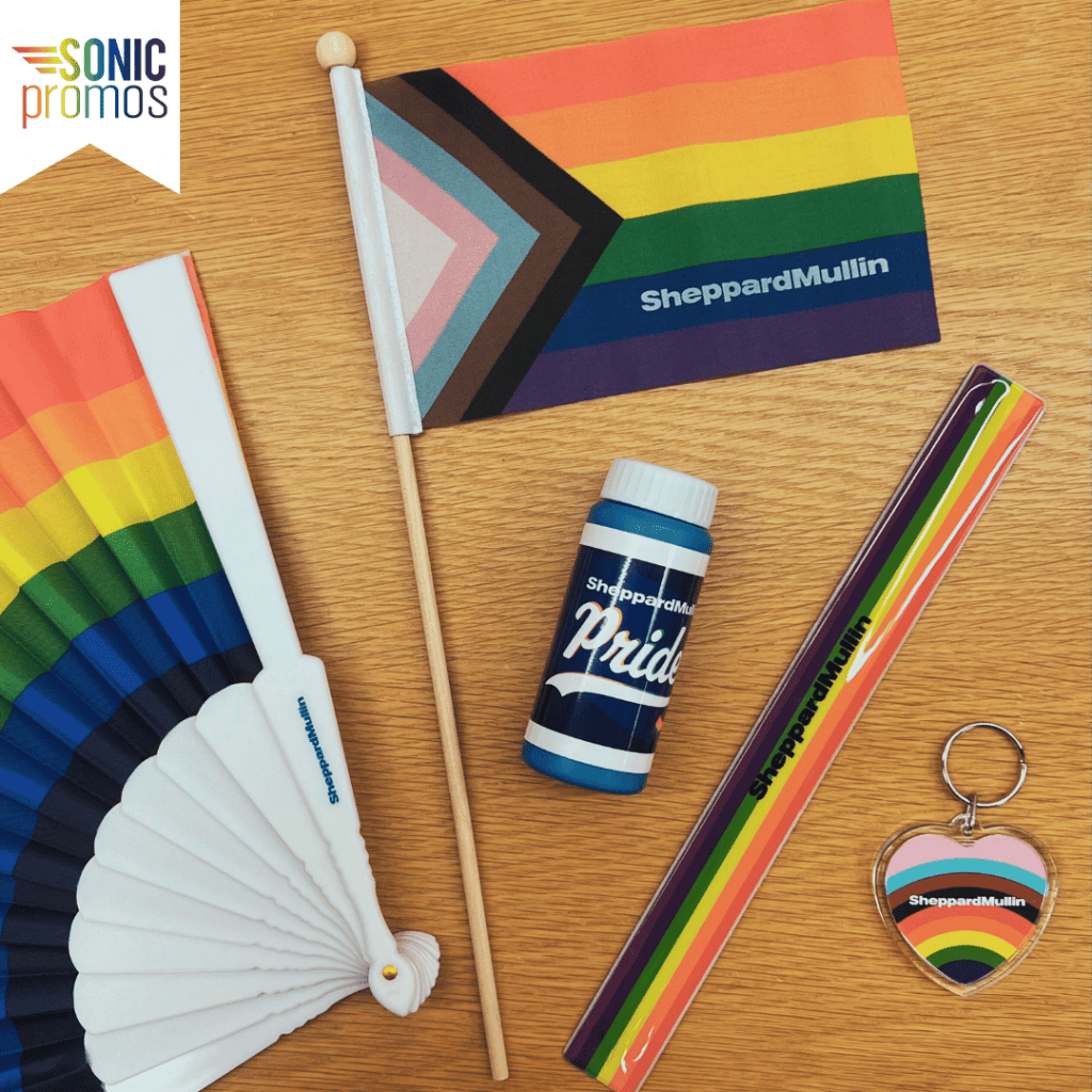 Custom branded Pride merch on a desk. Each is branded with the Sheppard Mullin logo: a rainbow fan, a rainbow Pride flag, bubbles, a slap bracelet and a keychain