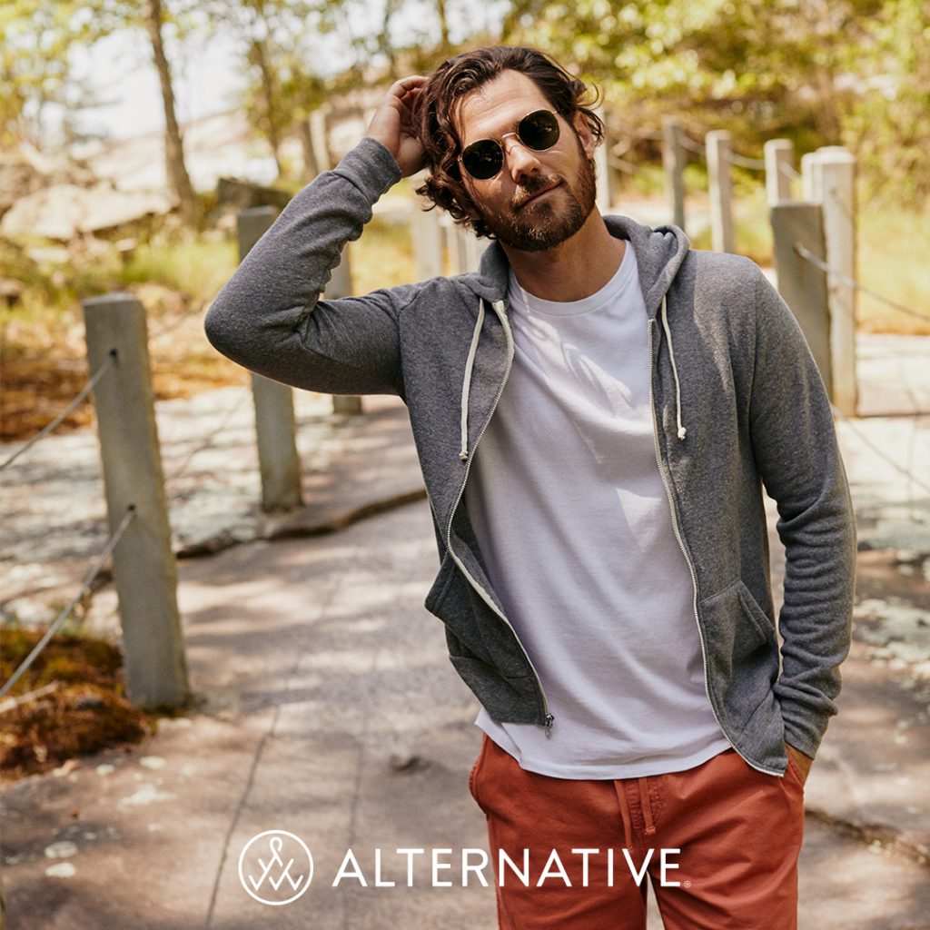 A man stands wearing sunglasses on a wood bridge in the woods. He's wearing a grey zippered hoodie that is unzipped and shows a shirt underneath it. He stands with one hand scratching his head and one hand in the pocket of his pants. The Alternative logo is at the bottom of the image