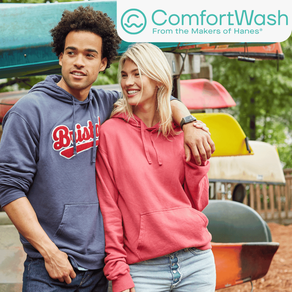 An adult man and woman stand with their arms around one another, smiling and looking off camera. The man stands to the left and is wearing a blue hoodie with some words printed on the front and the sleeves rolled up. The woman is wearing a pink hoodie. The background shows canoes stacked on a campground. The ComfortWash from the Makers of Hanes logo is in the top right corner.