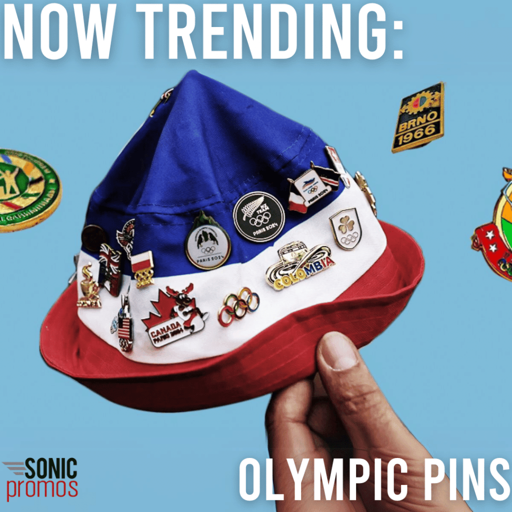 A blue, white and red striped bucket hat is being held against a blue backdrop. The hat is decorated with custom Olympic trading pins. Text reads: "now trending: Olympic pins."