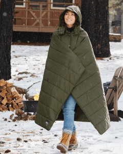 A woman is walking in a snow covered landscape, wearing a large military green Eddie Bauer poncho. She has the hood up