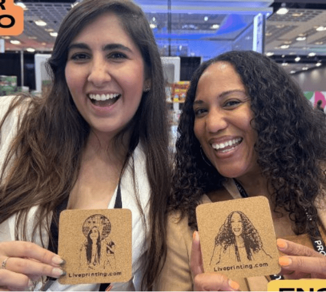 Two smiling women at an exhibition booth holding cork coasters with the 'Liveprinting.com' logo.