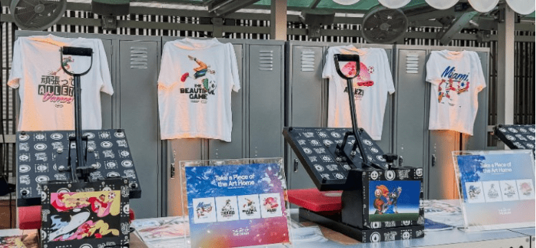 Row of white graphic tees hung on gray lockers at a booth, with display units and colorful art panels below.